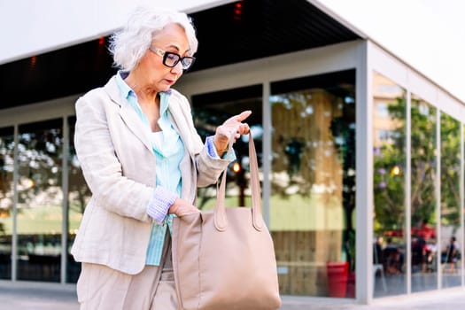 stylish mature woman looking in her bag while walking next to a building, concept of urban lifestyle in adulthood, copyspace for text