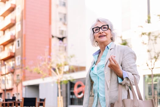 stylish mature woman with bag hanging on arm walking through the city while smiling happy, concept of urban lifestyle in adulthood, copyspace for text