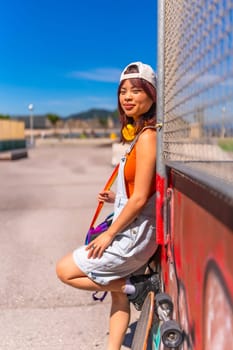 Vertical side view portrait of a young asian female skater leaning on an urban wall smiling at camera