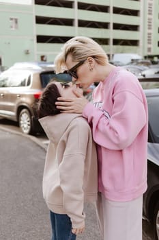 Candid moment between a mother and her daughter standing in a parking lot. Both are dressed in casual, fashionable outfits with sunglasses, emphasizing a relaxed and stylish urban lifestyle.