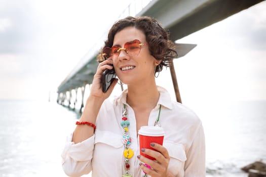 Woman in sunglasses on vacation on the beach, talking on mobile phone and drinking coffee