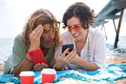 Middle-aged man and woman sitting on the beach with surprised face, browsing smartphone apps. Concept:Holidays y Technology