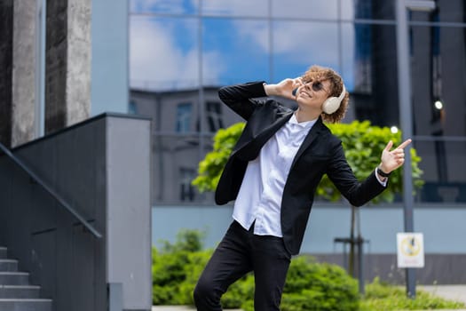 Happy relaxed young Caucasian businessman in wireless headphones dancing, listening favorite energetic disco music dancing outdoors. Overjoyed guy in suit outside office building in downtown city.
