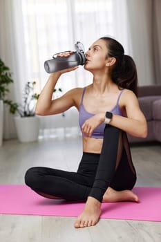 athletic fitness caucasian woman drinking water sitting on pink exercise mat at home