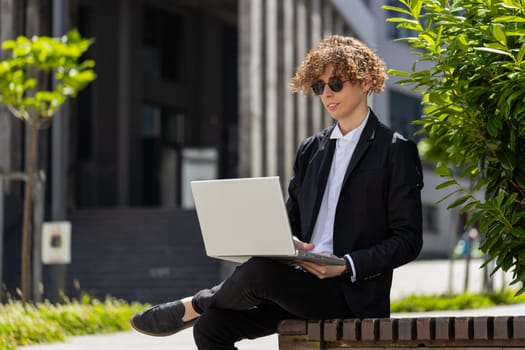 Happy smiling positive Caucasian businessman investor employer entrepreneur success with online project typing laptop outdoors in city. Positive freelancer guy in suit sitting against office building.