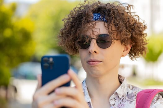 Caucasian young man using smartphone typing texting social media messages e-mail looking for a way on map in mobile navigator app outdoors. Smiling guy with backpack on city street. Town lifestyles.