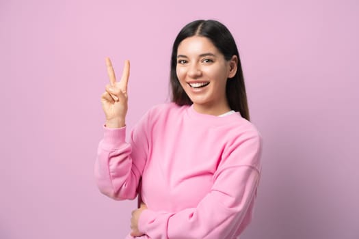 Vibes on. Young woman rocking a pink pullover, flashing a peace sign, and grinning ear to ear. Pink background for max pop. Perfect for modern, fun, and positive vibes.