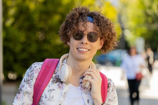 Portrait of happy smiling Caucasian young man listening music taking off headphones looking at camera, relaxation feel satisfied good news outdoors. Positive guy with curly hair on urban city street.