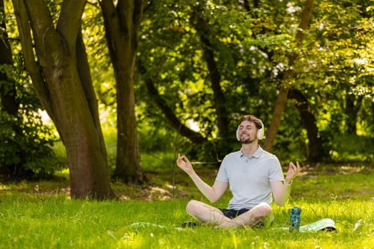 Young man sitting on mat in lotus position, relaxing in city park on summer day. Sporty carefree guy practicing yoga meditation, receives positive energy, healthy lifestyle. Active relax sportsman.