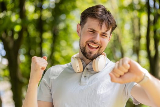 Happy active male runner jogging in city park and looking at smartwatch. Excited man using fitness tracker and checking result celebrating success. Athlete guy is satisfied with results of training.