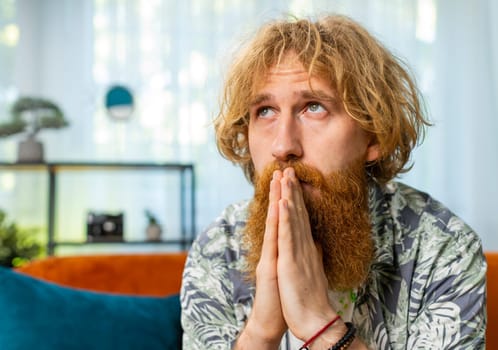 Please, God help. Worried Caucasian redhead man praying sincerely with folded arms, looking upward and making wish, asking with hopeful imploring expression, begging apology. Young bearded guy at home