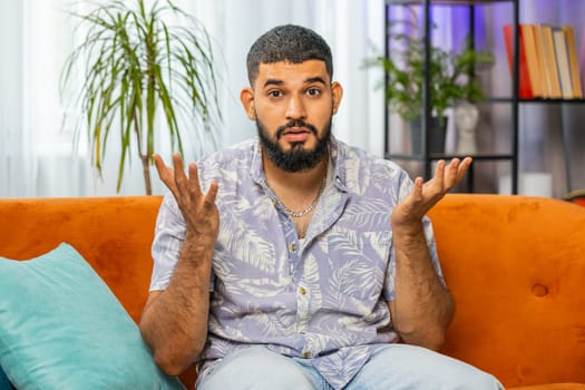 What. Why. Frustrated Indian man raising hands in indignant expression, asking reason of failure, demonstrating disbelief irritation by troubles, quarrel at home room apartment. Guy sits on couch