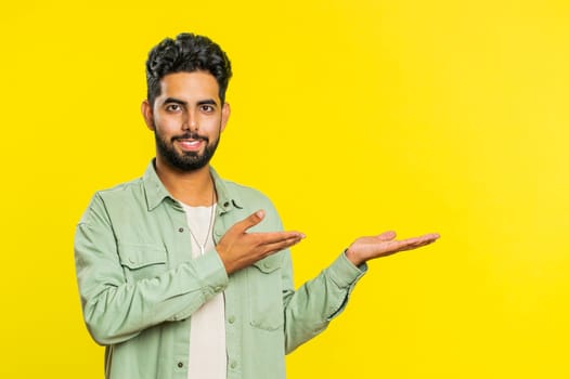 Indian bearded man showing thumbs up and pointing right empty place, advertising area for commercial text copy space for goods promotion advertisement. Guy isolated on yellow background. Lifestyles
