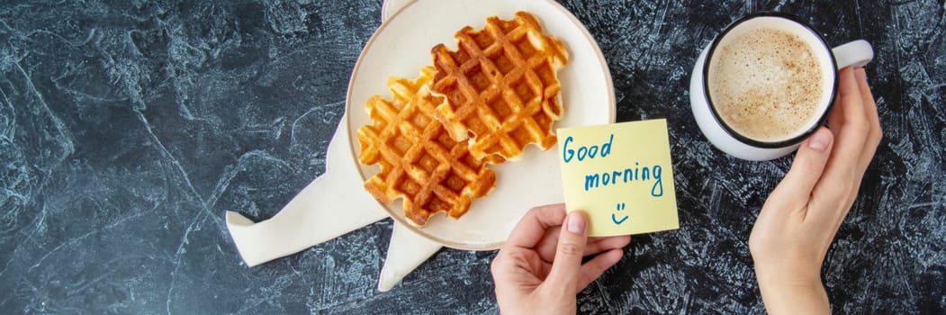 Banner of womans hands holding a note with the words GOOD MORNING next to a cup of coffee and spotted homemade Viennese waffles on a black textured background. breakfast flat lay. top view
