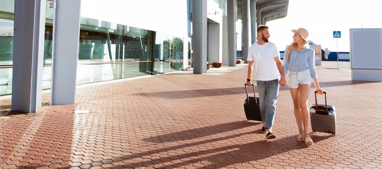 Travel Concept. Young couple walking with luggage near airport building, holding hands, empty space, full length