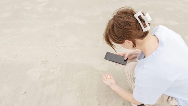 A young woman with beautiful long brown hair, in a blue shirt, joyfully crouches on the sandy shore, engrossed in her smartphone, sharing moments with loved ones at the beach.