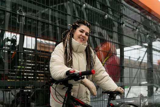 A woman with dreadlocks is standing next to a bicycle, looking at something in the distance. She has a relaxed posture, and the bicycle is parked on a city street.