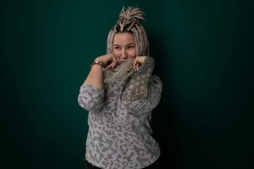 A woman with dreadlocks is standing upright against a bright green, solid wall. Her posture is assertive, looking directly ahead. The contrast between her dark hair and the vivid green background is striking.