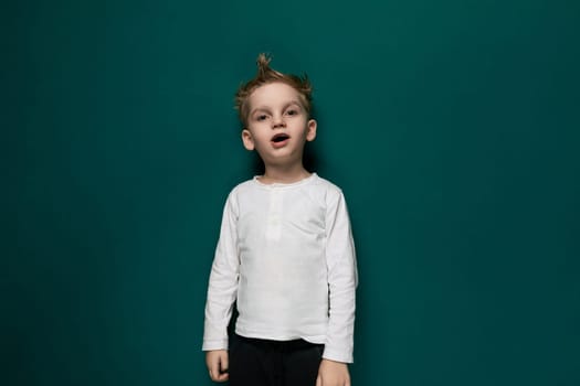 A young boy is standing in front of a solid green wall, looking directly at the camera with a neutral expression. The wall serves as a simple backdrop, highlighting the boys presence in the scene.