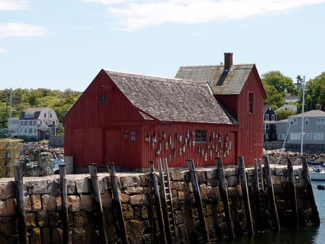 Rockport - June, 6 2024: A red wooden building stands on a stone pier in Rockport, with a small boat in the water.