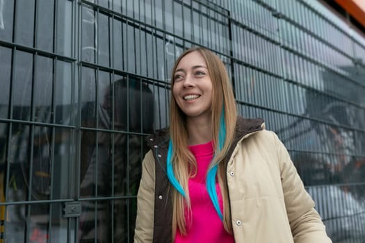 A woman is standing upright in front of a metal fence, looking straight ahead. She appears calm and composed, with no particular expression on her face. The metal fence is made of vertical bars and presents a barrier between the woman and what lies beyond.