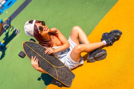 Cool young woman in street clothes, cap and sunglasses sitting on colorful urban skateboard park