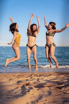 Three happy girls in jumping motion with on the beach outdoors in summer celebrating vacation days. Friends having fun on vacation by the sea carefree, and party concept