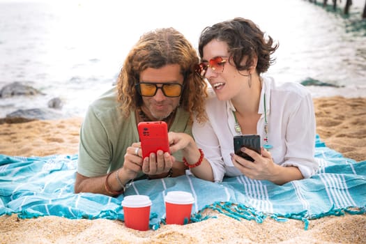 Middle-aged man and woman sitting on the beach with happy face, browsing smartphone apps. Concept:Holidays y Technology