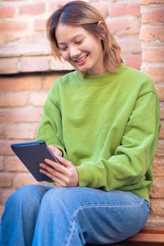 Vertical portrait beautiful Chinese teen student smiling using social media app on digital tablet outdoors. Girl sitting chatting on digital tablet