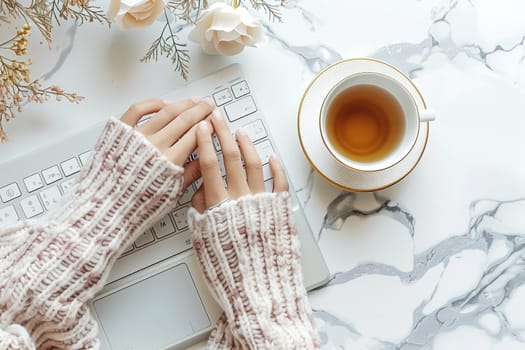 A person's hands, wearing a cream-colored knitted sweater, are typing on a laptop keyboard. A cup of tea and a saucer are to the right of the keyboard, and a few white and pink flowers are in the background.