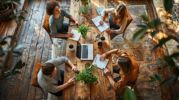 Four people sit around a large wooden table in a modern office, engaged in conversation and working on projects. They have a laptop, notebooks, and writing utensils on the table, surrounded by potted plants. The room has hardwood floors and the overhead view gives an intimate perspective on their collaboration.