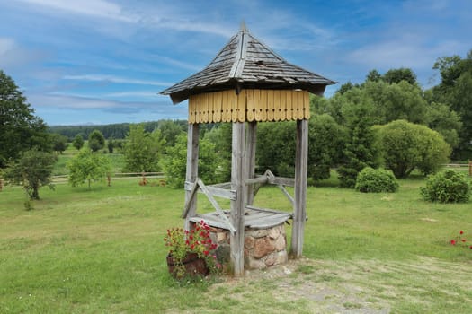 The well is made of stone and wood, the base is made of small stones. The roof is wooden, against the background of green grass, forest, sky.