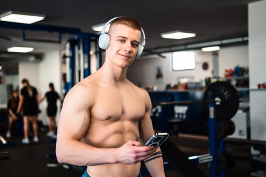 Portrait of a bodybuilder in a gym wears headphones and looks at his phone while resting