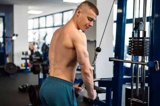 Back view of young man performs a triceps exercise with a cable machine at the gym