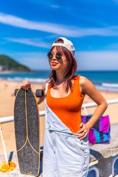 Vertical portrait of a young asian distracted woman with skateboard standing next to the beach