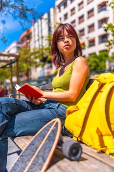 Vertical photo of a chinese young woman writing notes on diary sitting on park bench next to skateboard