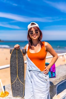 Vertical Portrait of a cool young female asian skater in summer modern clothes, cap and sunglasses smiling at camera standing next to the beach