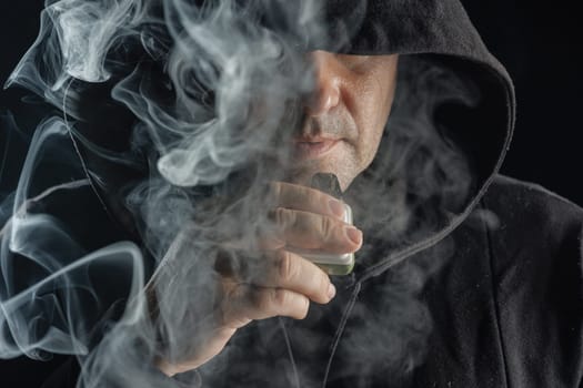 a man smokes a vape electronic cigarette in black clothes on a dark background, close-up, nicotine addiction, Harm of electronic cigarettes