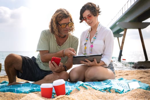 Middle-aged man and woman sitting on the beach with surprised face, browsing smartphone and tablet apps. Concept: Holidays and Technology