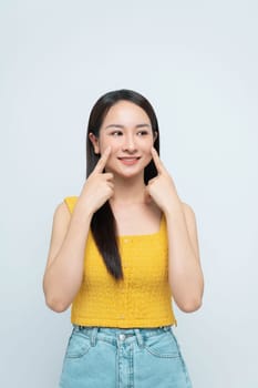 Cheerful woman pointing to her cheeks, expressing happiness and positivity on a light background.