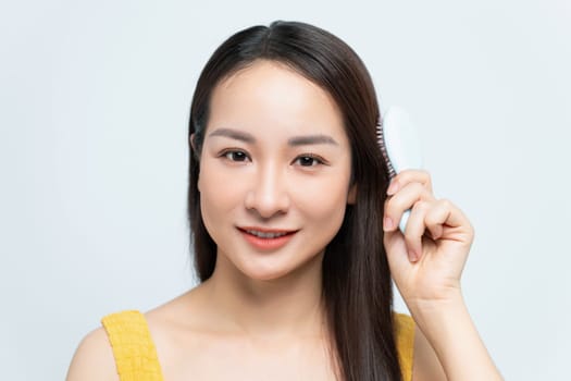 Smiling woman brushing her hair with a white hairbrush, wearing a yellow top and blue jeans.