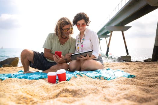 Middle aged man and woman sitting on the beach looking surprised, browsing smartphone and tablet apps looking at camera Concept:Vacation and Technology