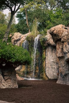 A picturesque waterfall flows in a natural wildlife sanctuary, surrounded by lush vegetation and rocky formations. The scene highlights the serene beauty and untouched nature of the environment.