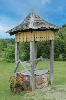 Wooden well water. The well is made of stone and wood, the base is made of small stones. The roof is wooden, against the background of green grass, forest, sky.