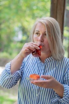 young blonde Caucasian woman drinks tea from Turkish Armudu cup on the balcony and takes bite of orange cake, delicious dessert after lunch, traditional oriental sweet on summer vacation