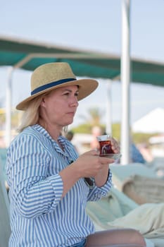 Young blonde drinks tea from Turkish Armudu cup,sitting on sun lounger on the sea beach, and looks at the sea, being in deep thought,wearing a long blue and white striped shirt and a straw hat