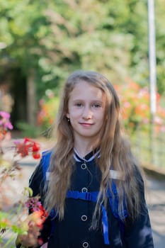 Back to school, portrait of a little girl from primary school outdoors, enjoying flowers in a flowerbed and looking at the camera with a smile, student dressed in dark blue uniform