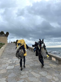 Back view anonymous underwater fisherman in wetsuits and gears walking near sea under cloudy sky. Rear view of male friends with spearguns and catch leaving after hunting.