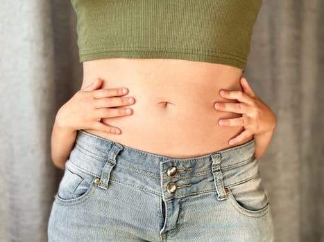 Close up of woman in jeans and green top with childs hands around her waist. Concept of family bonding, motherhood, postpartum recovery and affection. Neutral background. High quality photo