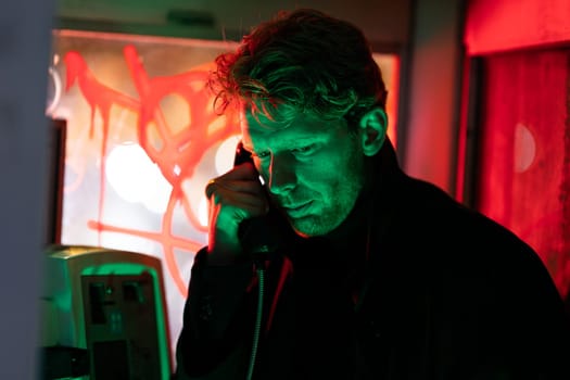 A man is talking on a phone at noir style in booth The image has a dark and moody atmosphere, with the man's face illuminated by the phone's light